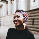 Smiling woman wearing vibrant headscarf and earrings, captured in an urban street setting.