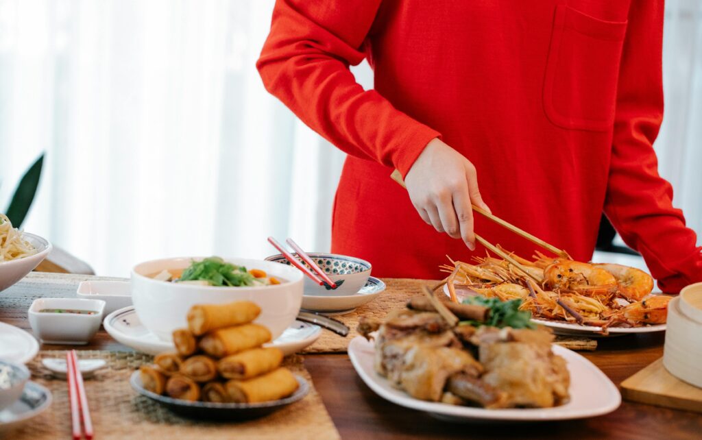 Crop unrecognizable female with tweezers serving tasty cooked prawns at table with spring rolls and stewed vegetables at home